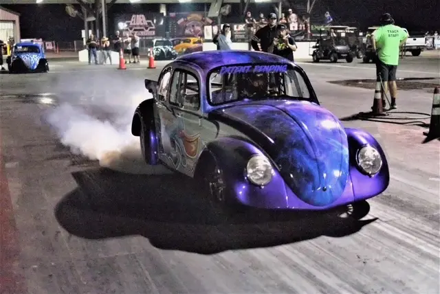 A classic purple Volkswagen Beetle racing at night, emitting smoke from its tires.